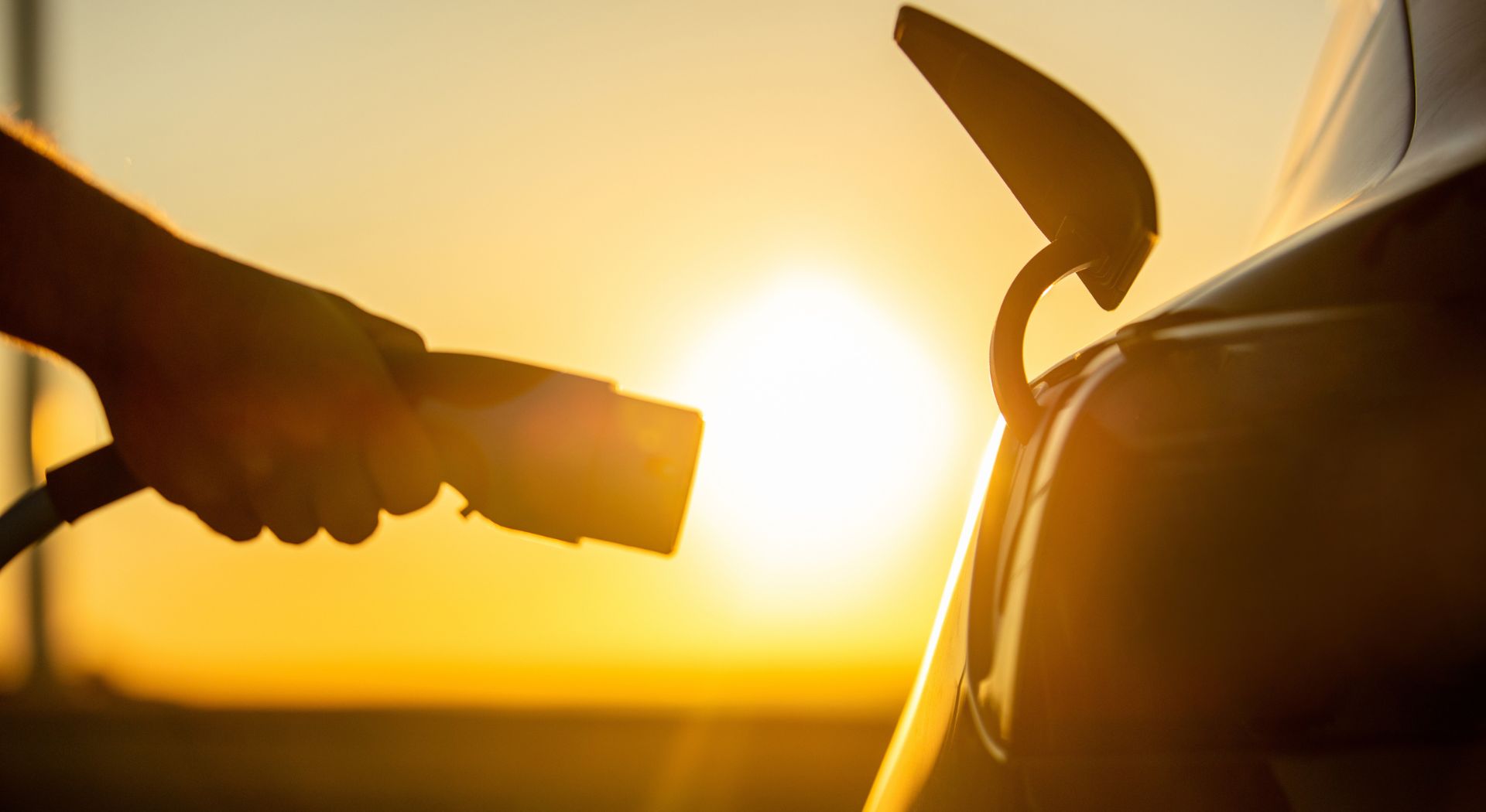 Electric vehicle user holding plug next to vehicle charging port, with sunset in background.